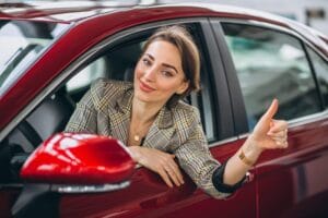 Mujer joven sonriente sacando el pulgar arriba desde la ventana de un carro rojo, en señal de aprobación o logro.